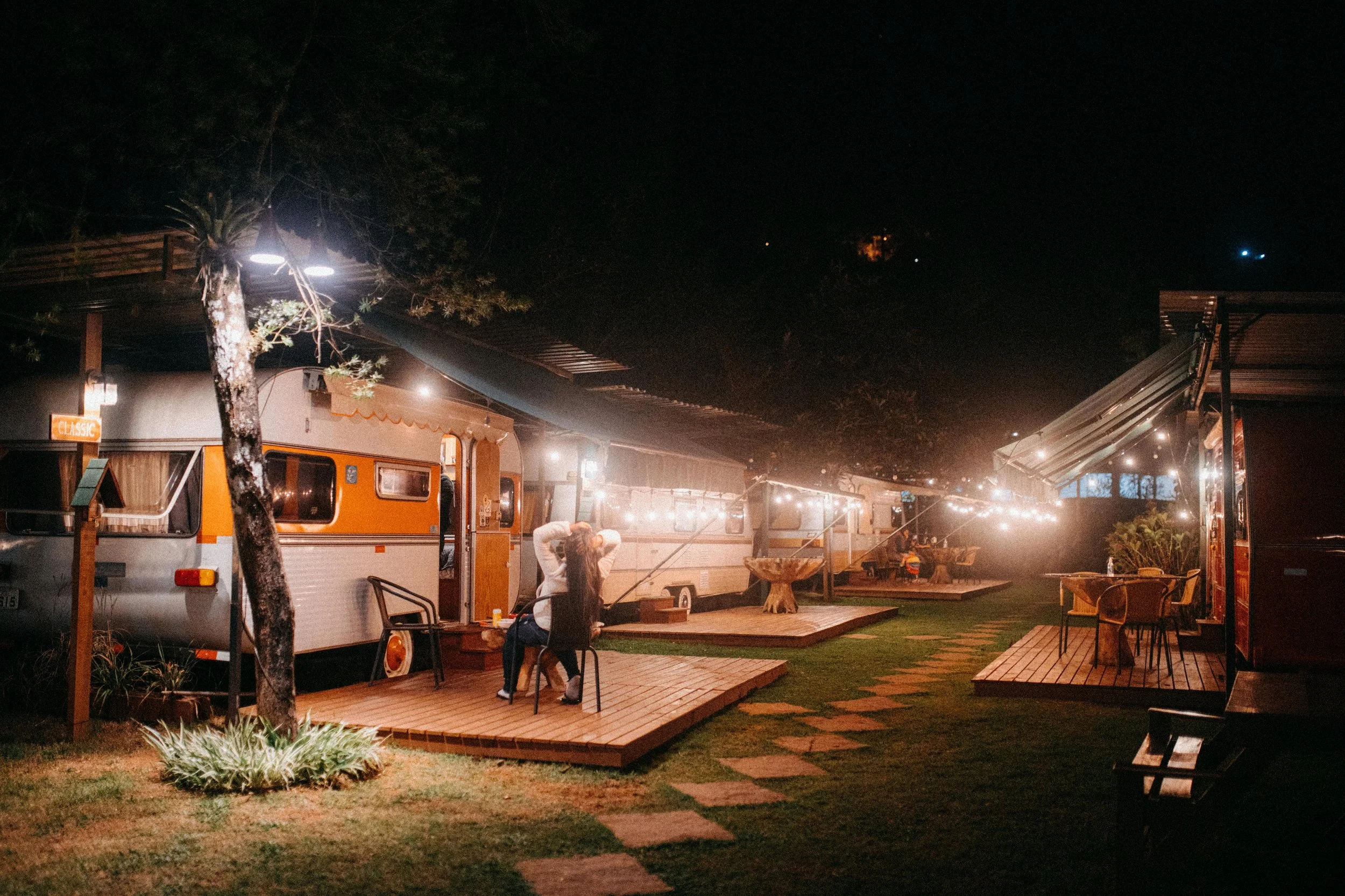 Night scene of outdoor seating area with a vintage camper trailer, string lights, cozy wooden decks, outdoor furniture, and a person sitting on a chair, surrounded by trees.