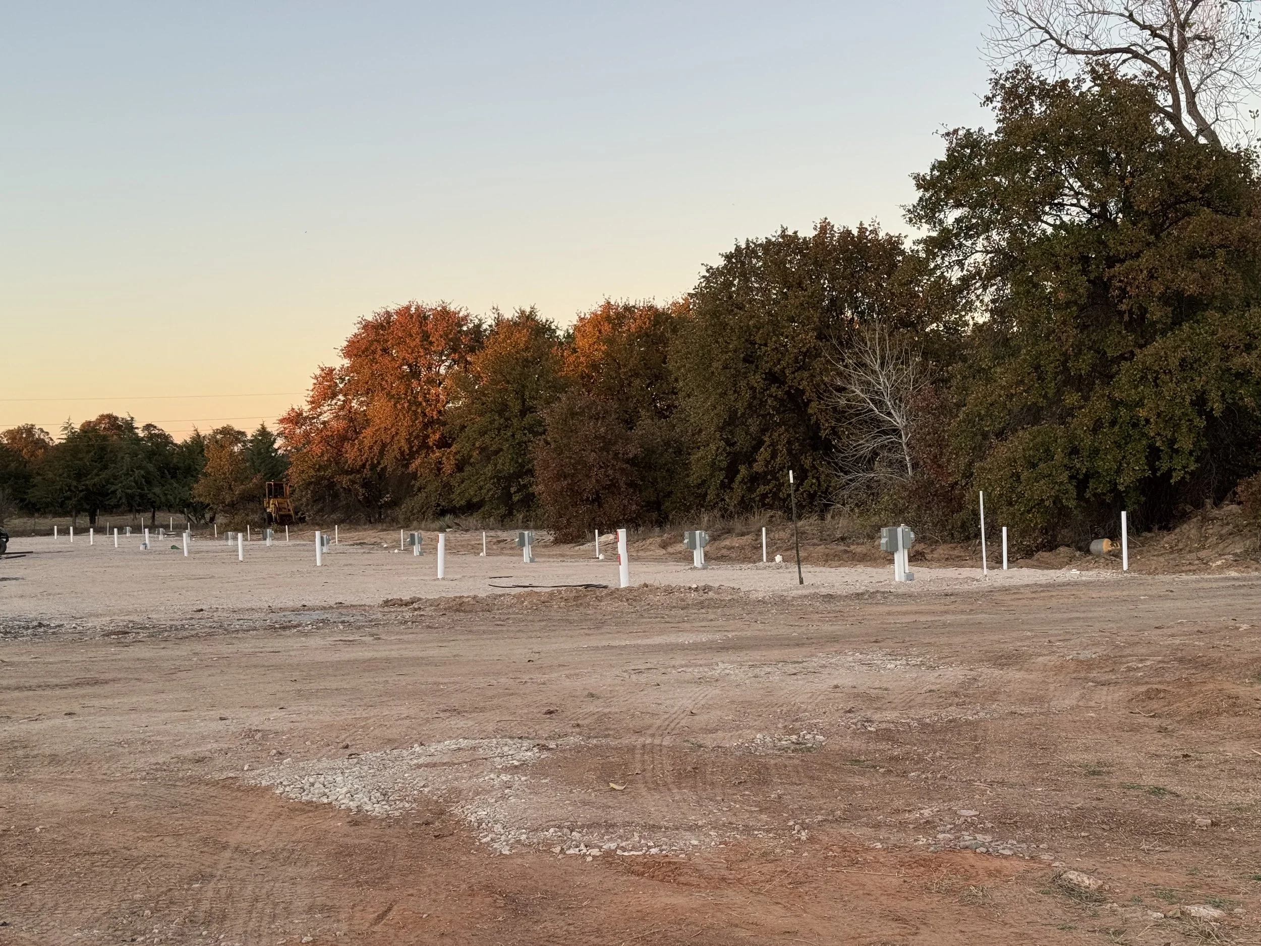 Construction site with utility boxes and poles, trees in the background, and a dirt ground under a clear sky.