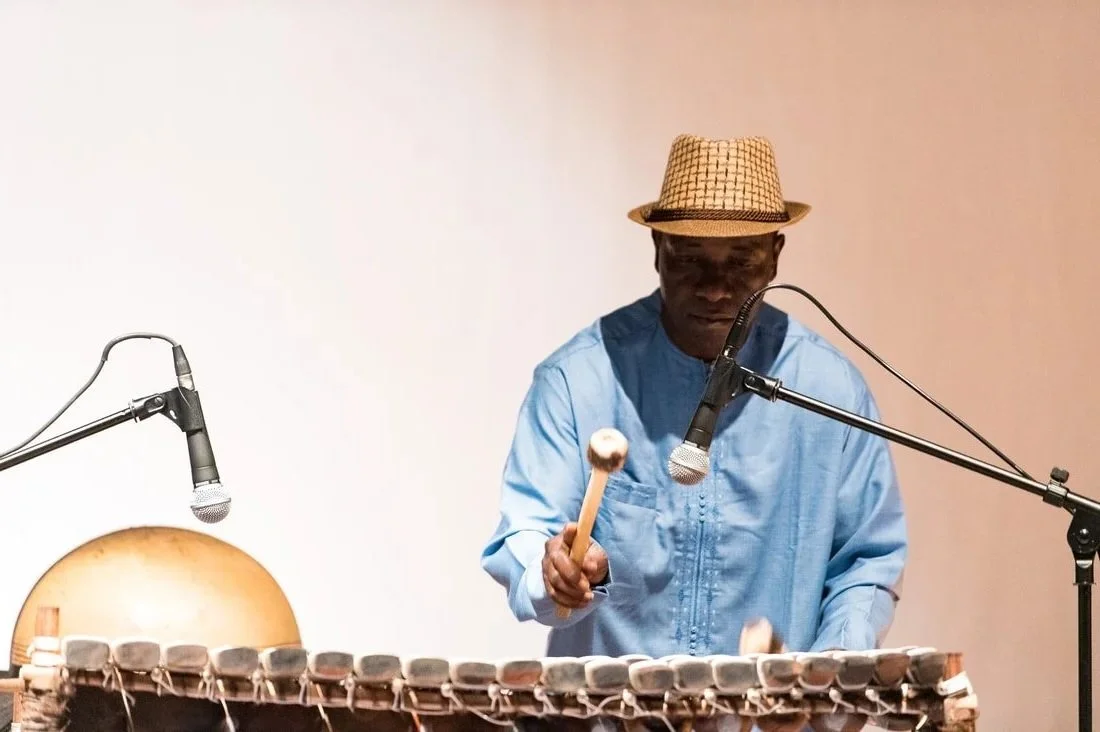 Man wearing a straw hat and blue shirt playing a xylophone with a mallet, surrounded by microphones.