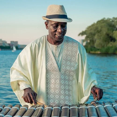 Man wearing traditional attire and a fedora hat, playing a Balafon outdoors near a water body.