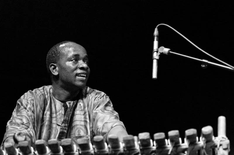 A Black man in traditional clothing speaking or performing at a sound mixing console with a microphone suspended nearby.