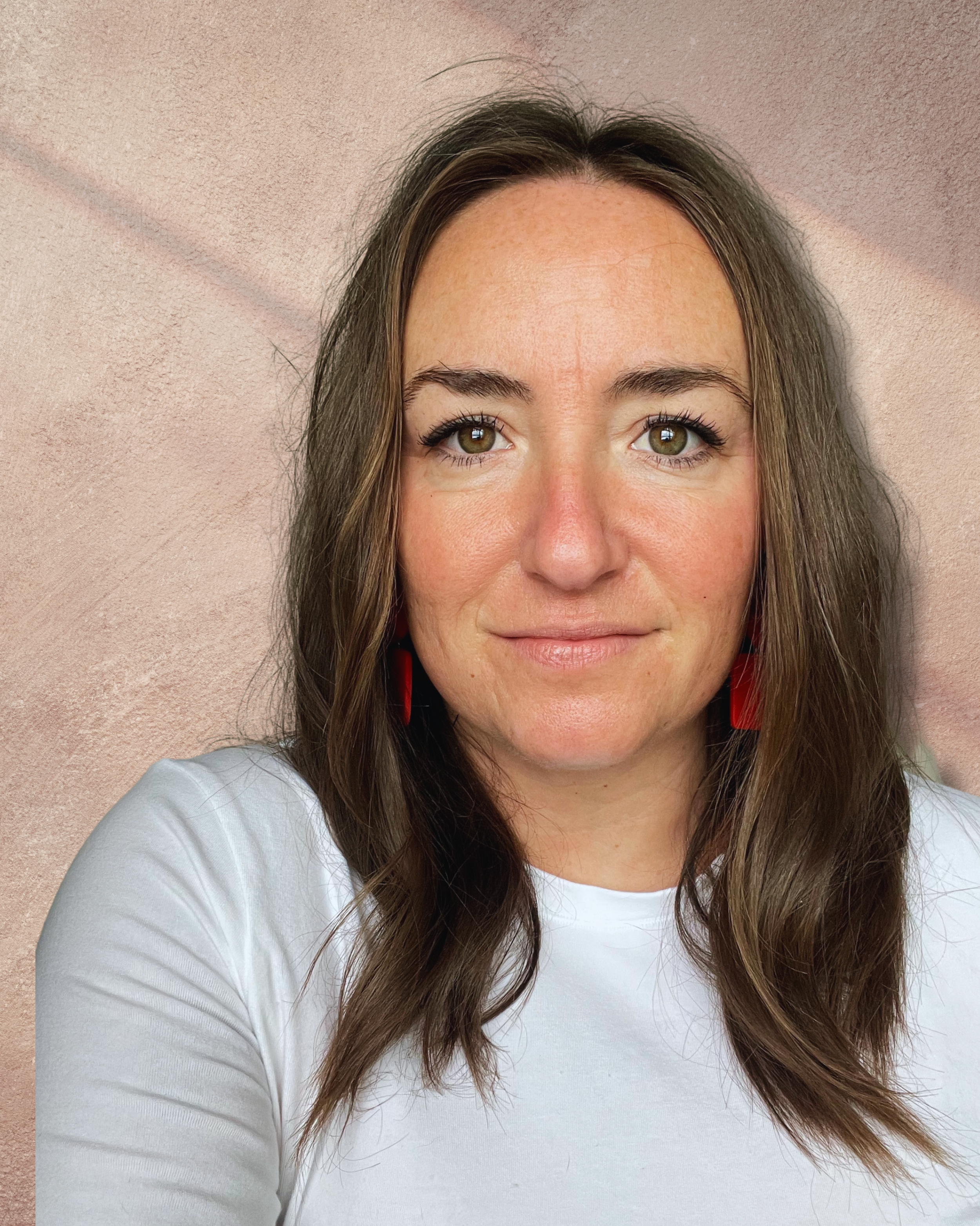 A woman with light skin, green eyes, and long wavy brown hair wearing a white shirt and red earrings, standing against a pink textured wall.