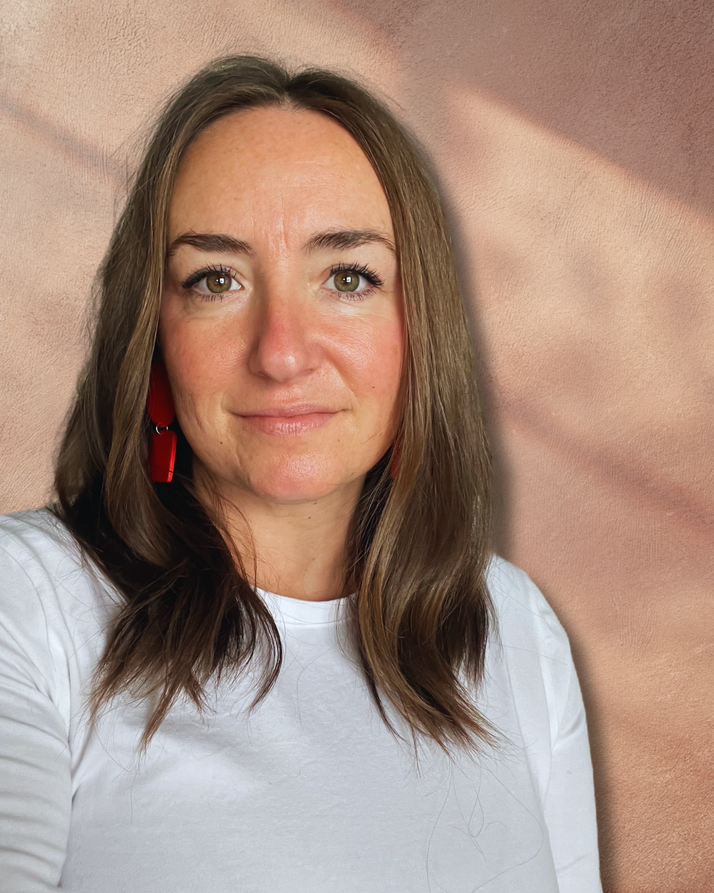 A woman with brown hair, wearing a white shirt and red earrings, smiling at the camera against a textured, peach-colored background.