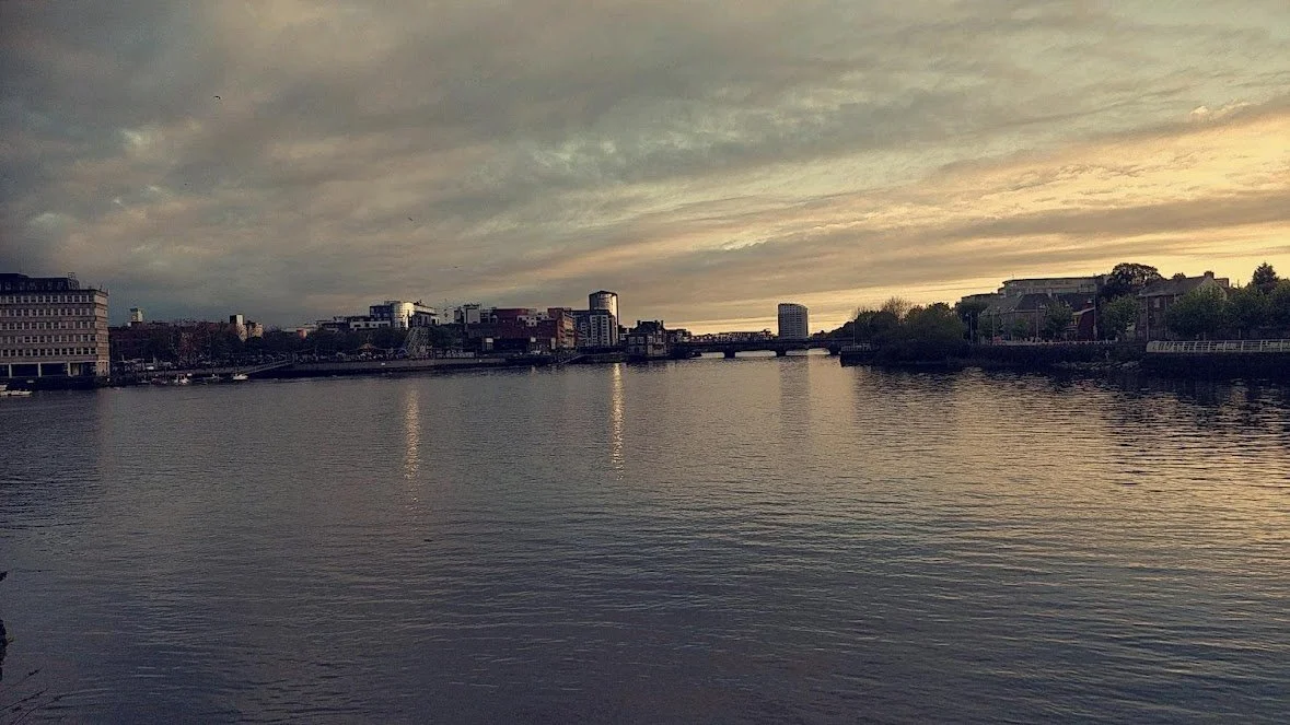A Photograph taken by Marlena Lambe showcasing a City skyline at sunset over a river with reflection of buildings and cloudy sky. This is Limerick City.
