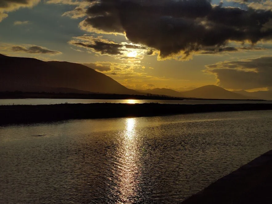 A Photograph taken by Marlena Lambe showcasing a Sunset over a lake with mountains in the background, partly cloudy sky, and the sun's reflection on the water. This is Cockleshell beach, Tralee.