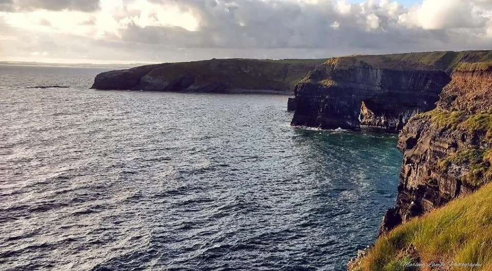 A Photograph taken by Marlena Lambe showcasing Cliffs in Ballybunion along the ocean with cloudy sky