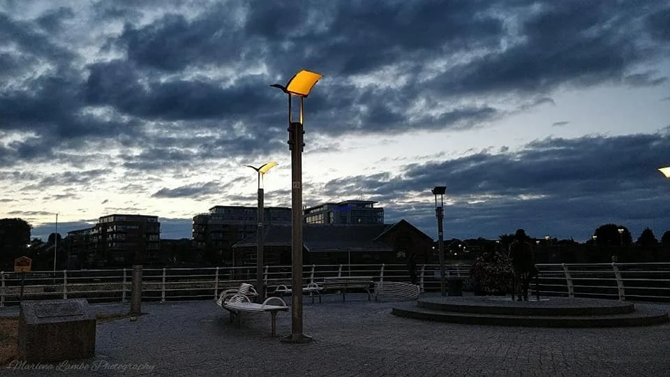 A Photograph taken by Marlena Lambe showcasing An outdoor urban park during early evening with benches, modern streetlights, and a person standing near a small pond or fountain, with overcast clouds in the sky. This was Limerick City, Ireland.