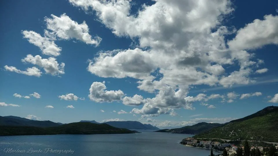 A Photograph taken by Marlena Lambe showcasing a scenic view of a river or lake with mountains on both sides and a partly cloudy sky. This was Neum.
