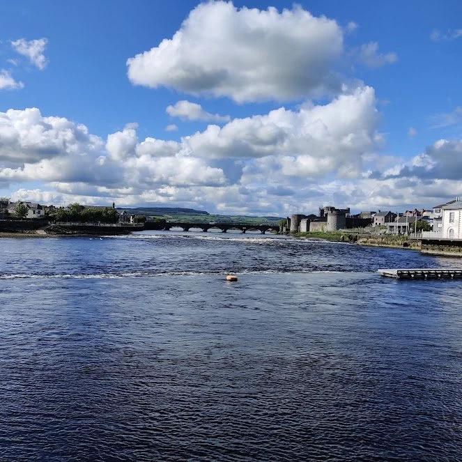 A Photograph taken by Marlena Lambe showcasing a Scenic view of a river with a stone bridge in the distance, buildings along the riverbank, under a partly cloudy sky. This is Limerick City.