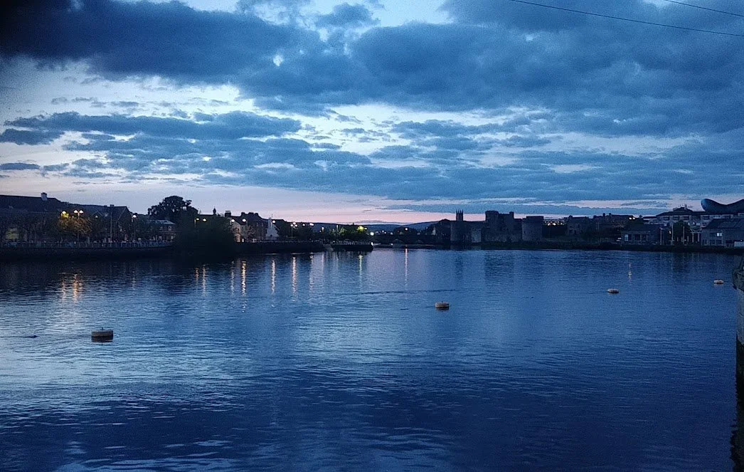 A Photograph taken by Marlena Lambe showcasing a Dusk over a river with calm water reflecting the cloudy sky, some buildings and a church in the distance, and a few buoys floating on the water. This is Limerick City.