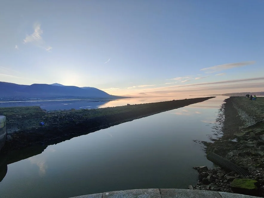 A Photograph taken by Marlena Lambe showcasing a calm river or canal reflecting the sky and a distant mountain range at sunset or sunrise, with a slightly cloudy sky and some people on the right side. This is Cockleshell beach, Tralee.