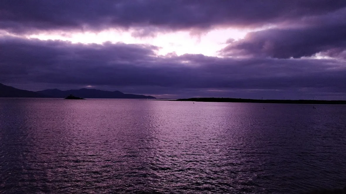 A Photograph taken by Marlena Lambe showcasing a body of water under a cloudy purple and gray sky, with distant mountains and a small island. This is Fenit, Kerry.