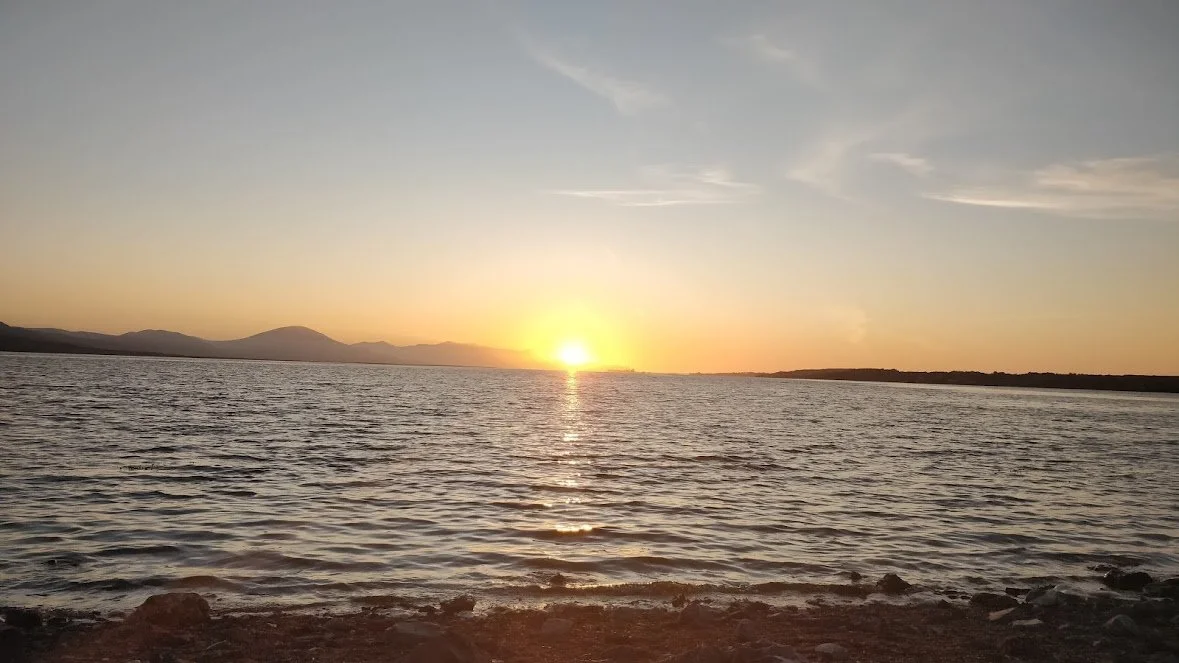 A Photograph taken by Marlena Lambe showcasing a Sunset over a body of water with a rocky shoreline in the foreground and mountains in the distance. This is Cockleshell beach, Tralee.