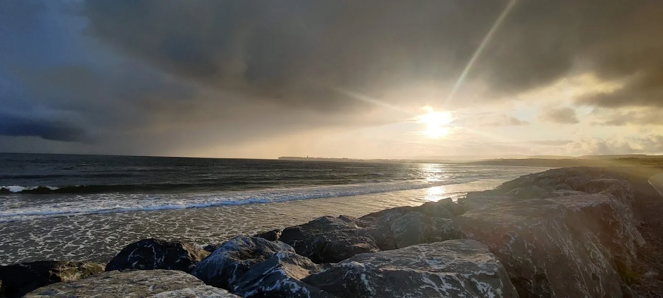 A Photograph taken by Marlena Lambe showcasing a Sunset over the ocean with waves crashing on the shore and large rocks in the foreground. This is Killkee.