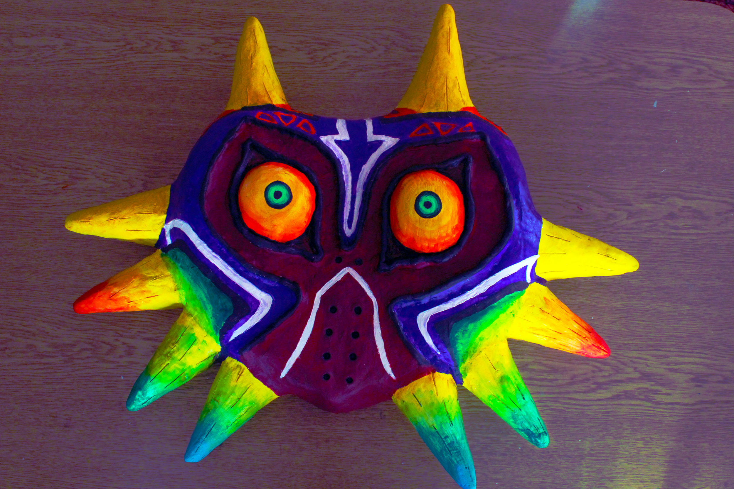 Colorful decorative mask with a face resembling Majora's Mask from a video game, featuring vibrant multiple colors including purple, yellow, orange, green, and blue, with large prominent eyes and spiked edges.