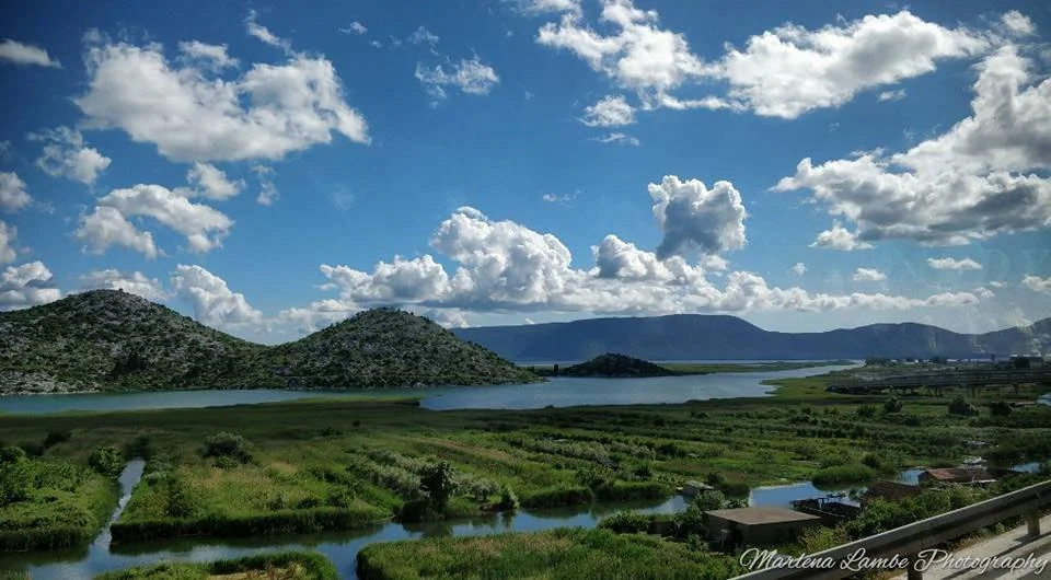 A Photograph taken by Marlena Lambe showcasing a Scenic landscape of green fields, water bodies, hills, and a partly cloudy sky. This was Neum.