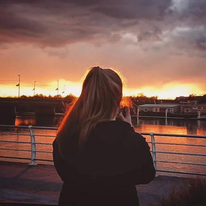 Marlena Lambe, A lady with blonde hair stands on a bridge, taking a photo of a sunset over a river, with dark clouds in the sky.