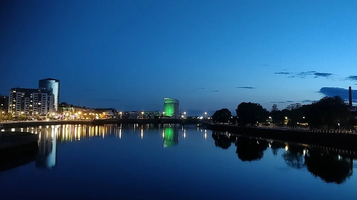 A Photograph taken by Marlena Lambe showcasing a Nighttime cityscape with tall buildings reflecting in a river, with a bridge in the background and trees along the riverbank. This is Limerick City.
