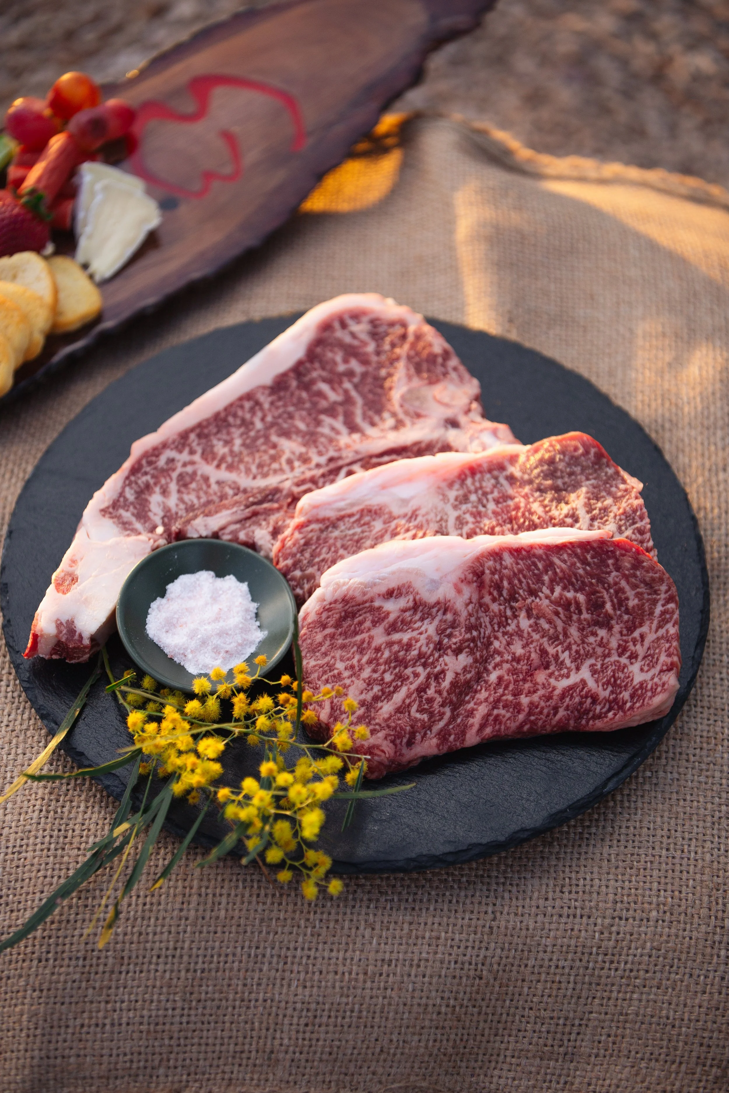 Three raw, marbled beef steaks on a black round slate plate, with a small dish of salt and yellow flowers, on a brown woven cloth.