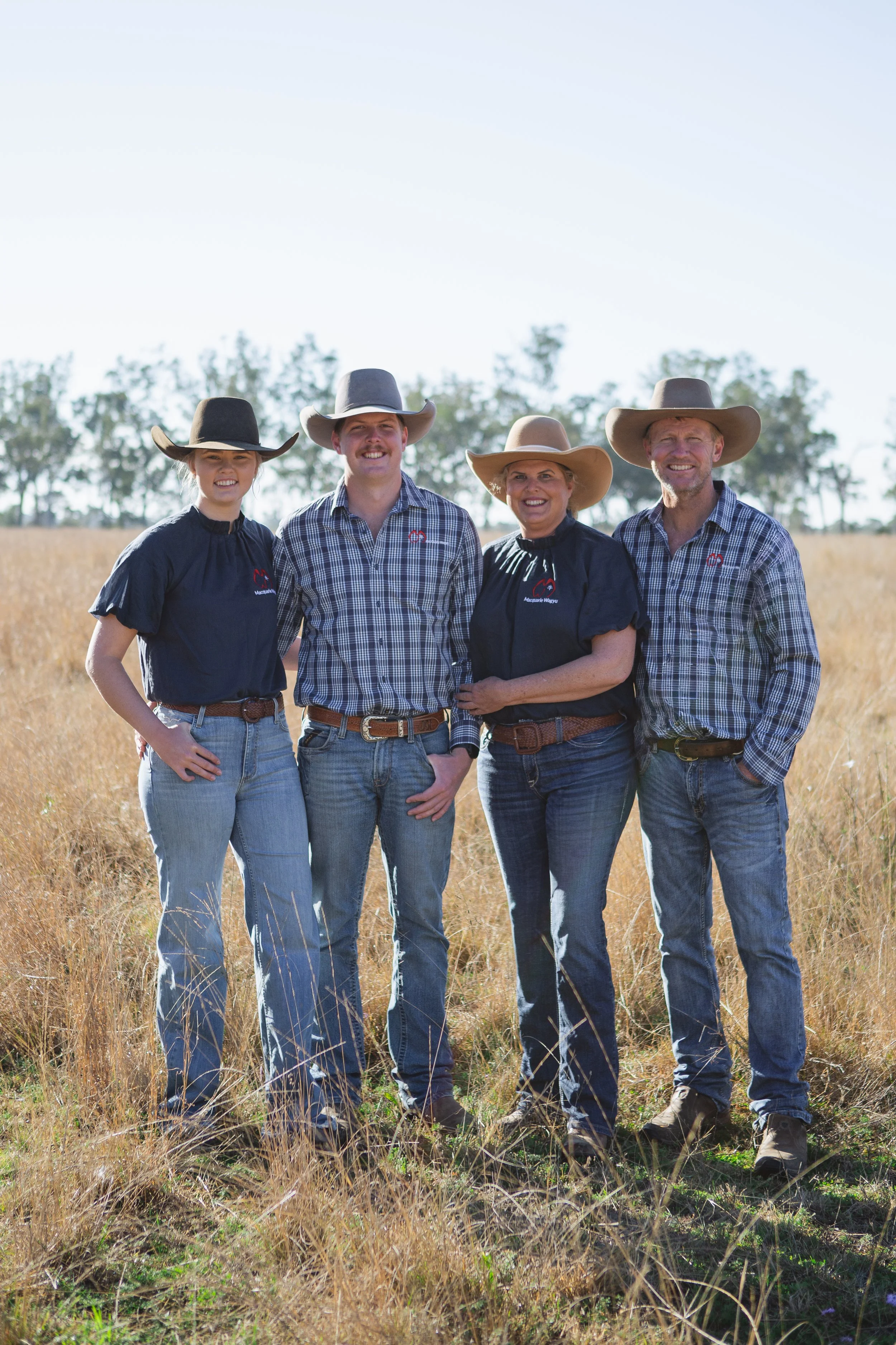 Family of four in cowboy hats and western attire standing in a grassy field with blue sky and trees in the background.