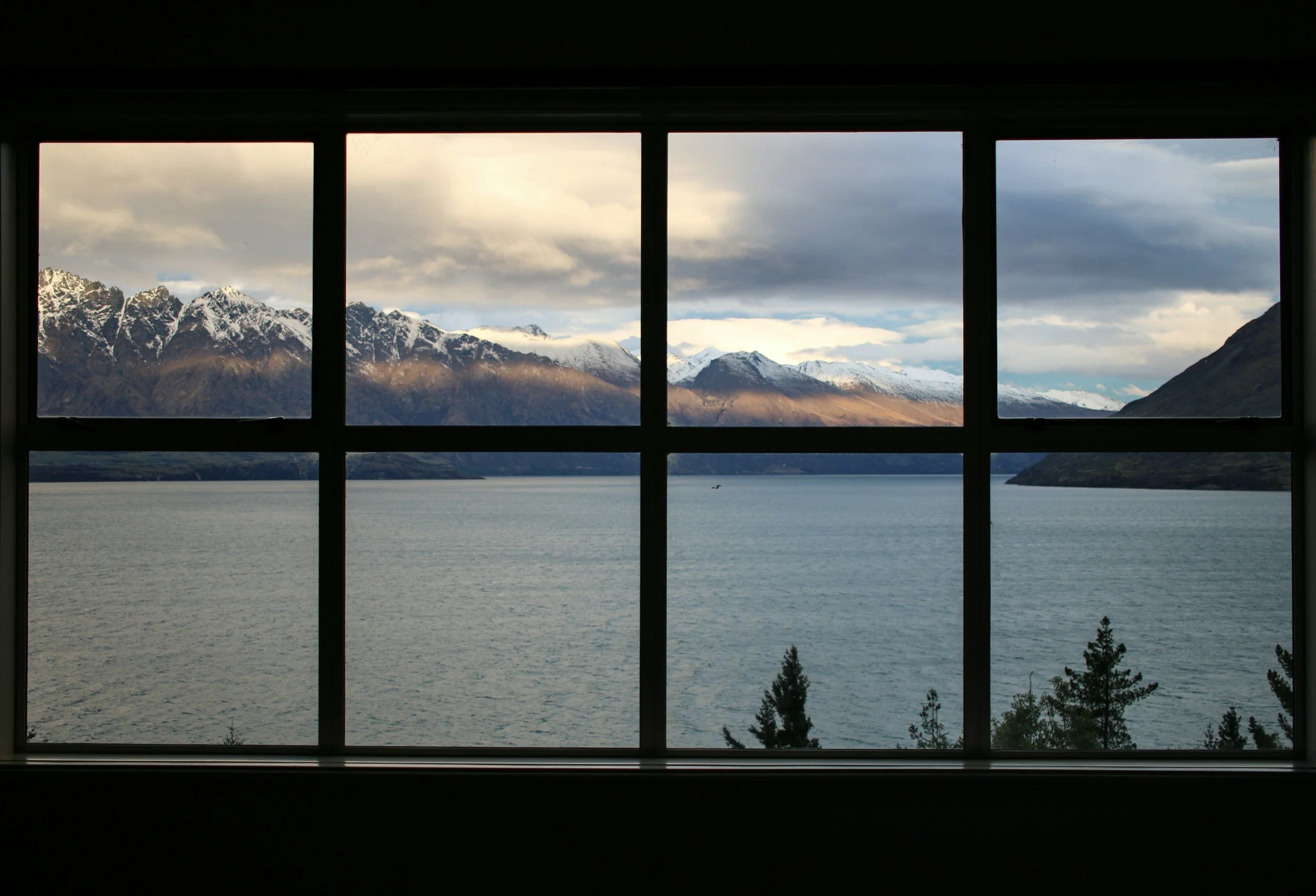 View of snow-capped mountains, cloudy sky, and a body of water seen through a window with black framing.