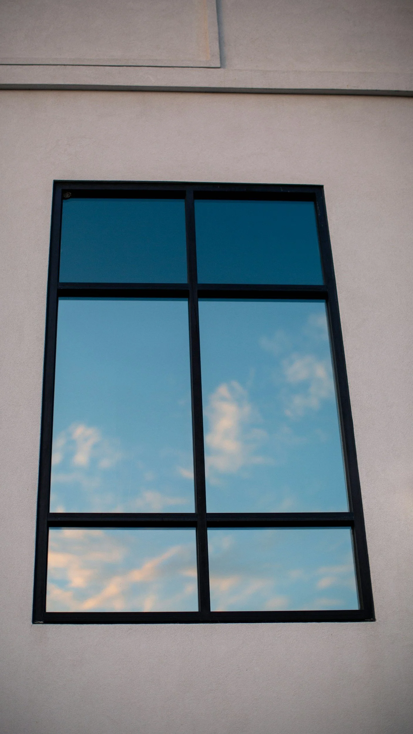 A window reflecting a blue sky with some clouds, on a building with beige exterior wall.