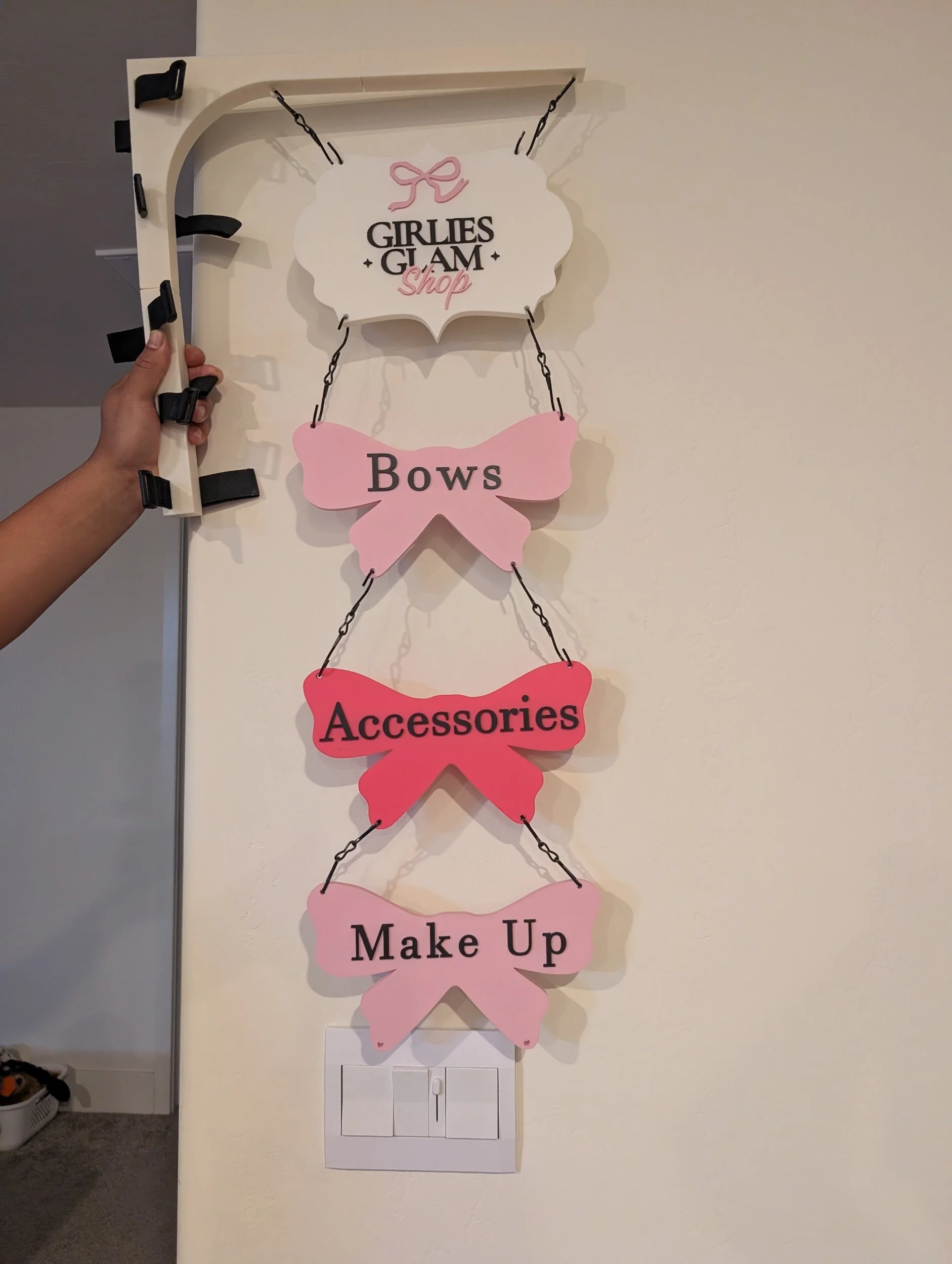 Decorative wall hanging with a white sign at the top that reads "Girlie's Glam Shop" with a pink bow above it, followed by pink paper bows labeled "Bows", "Accessories", and "Make Up" with chains connecting each bow, hanging on a wall.