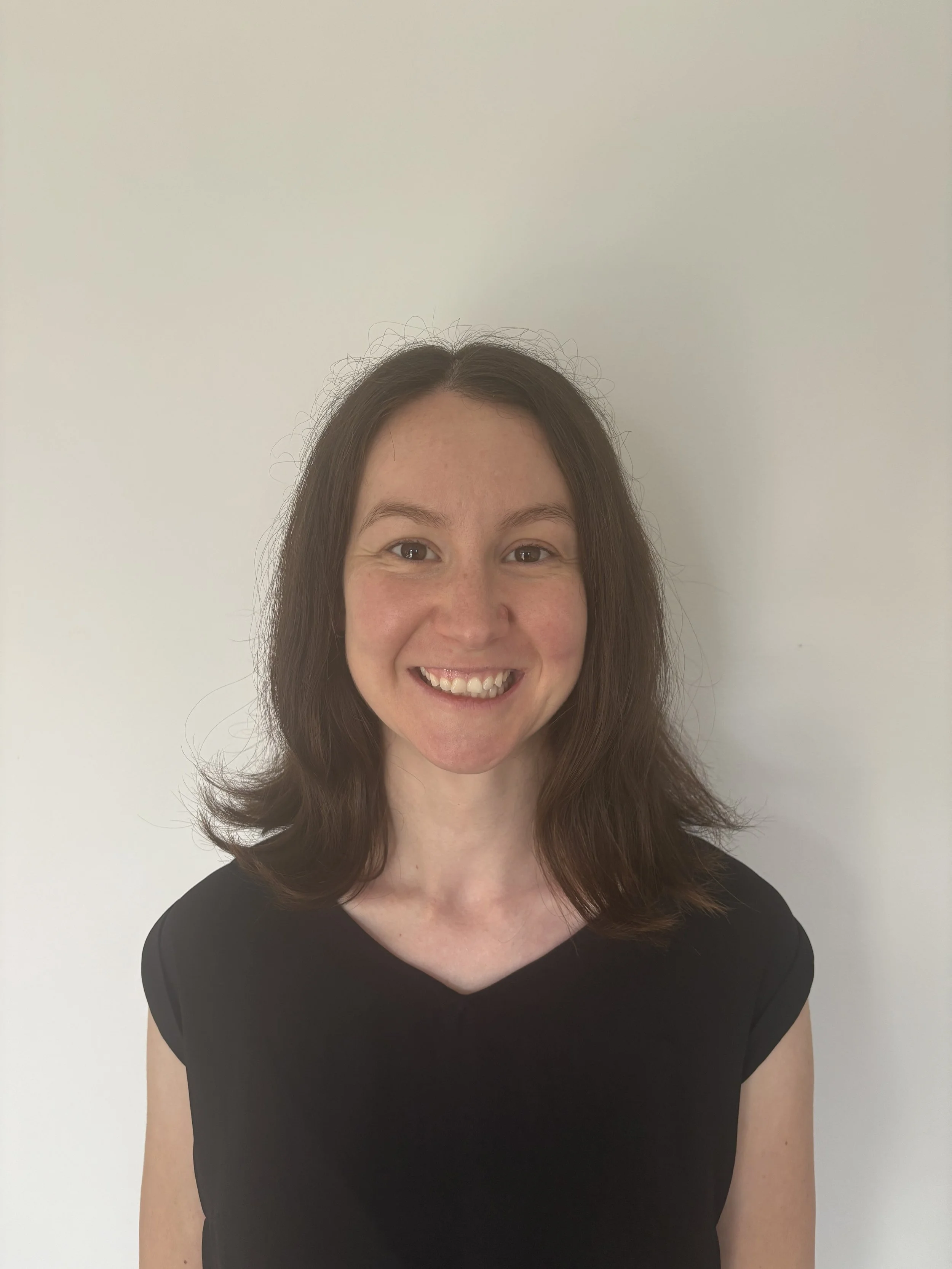 A woman with shoulder-length brown hair, smiling, wearing a black top, standing against a plain white wall.