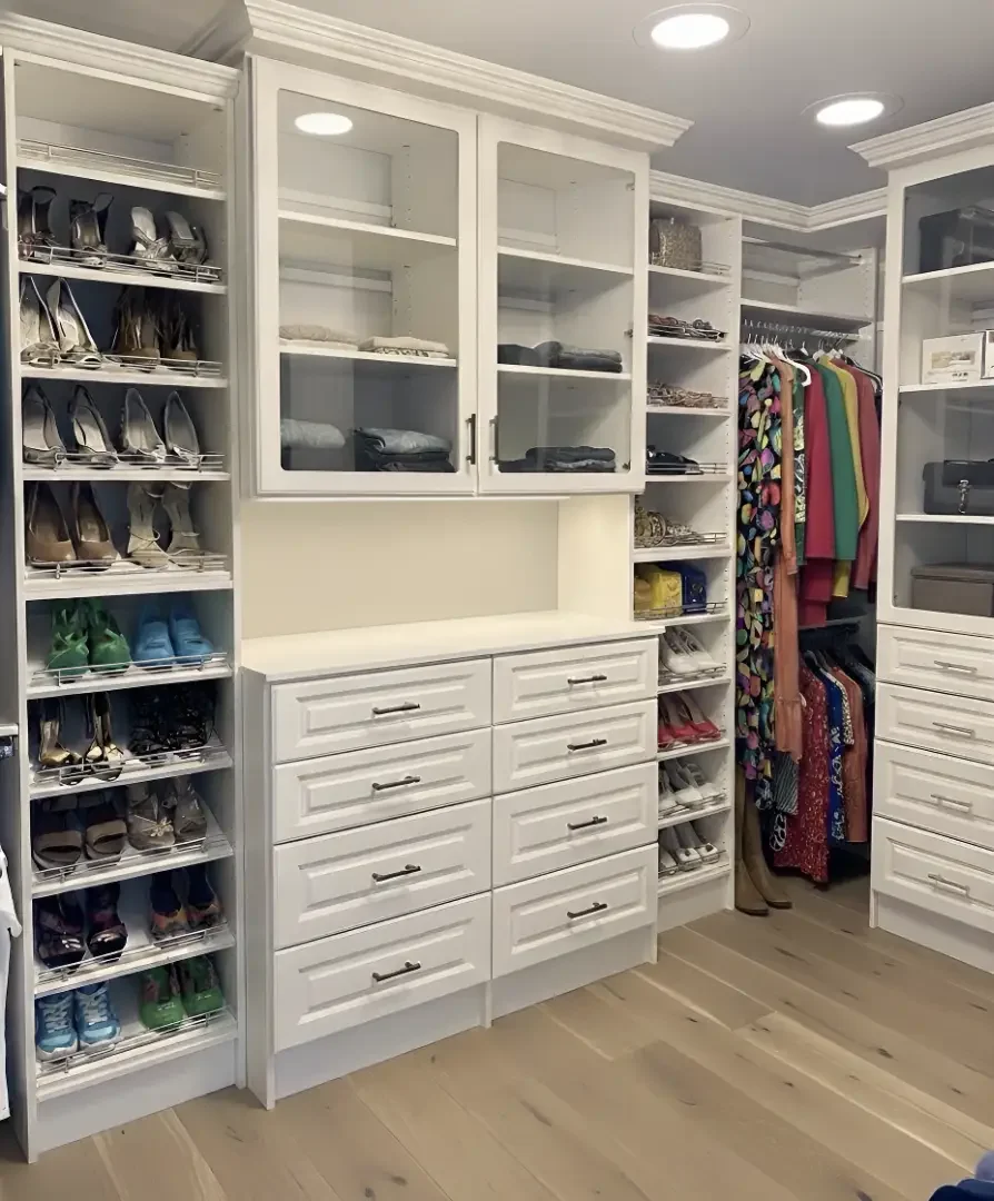 A walk-in closet with white wooden cabinets and shelves, containing folded clothes, shoes, and handbags, with hanging clothes to the right.