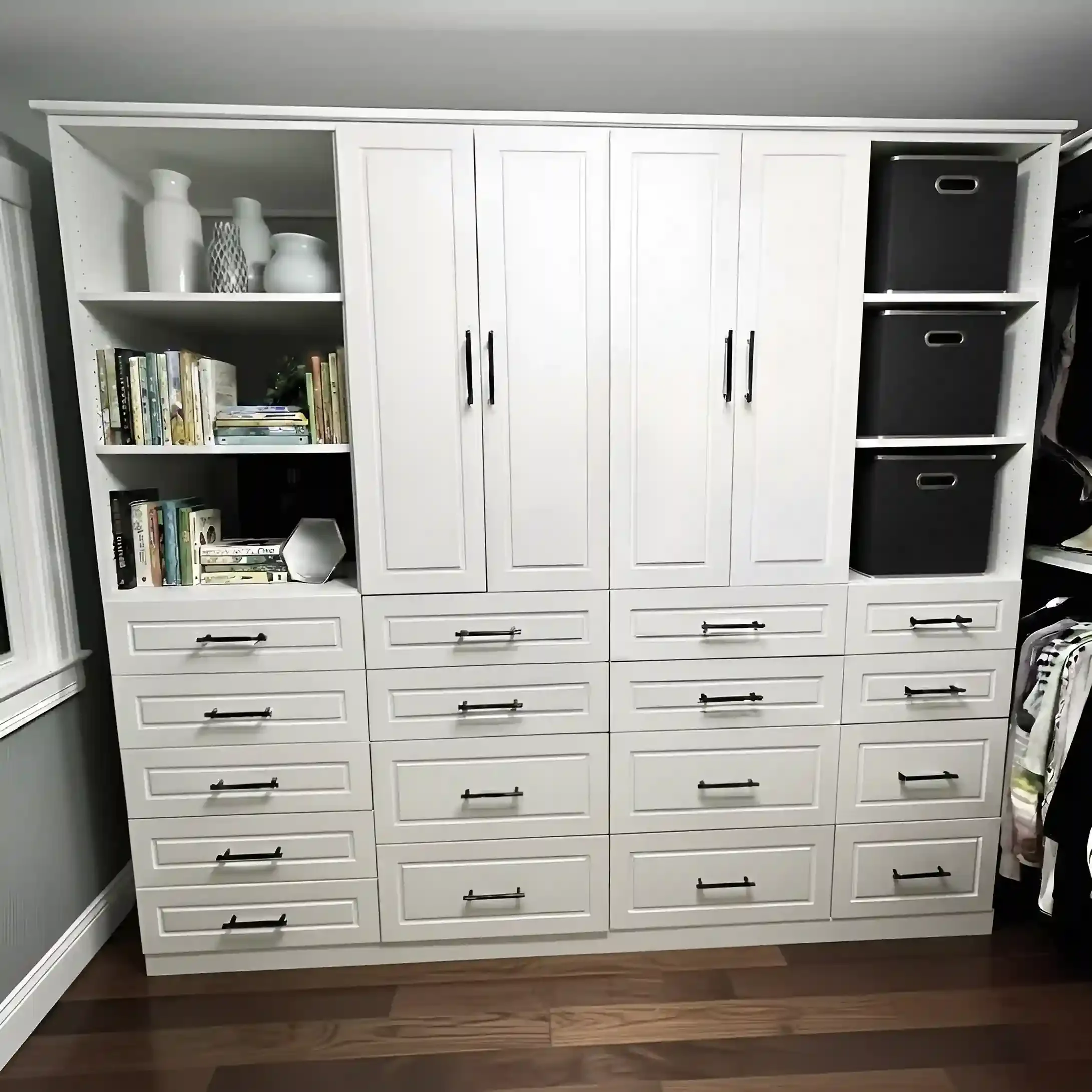 A large white storage cabinet with multiple drawers and compartments, featuring black handles. The top left shelves hold decorative vases and a collection of books, while three black storage bins are on the right side in a shelf compartment. The cabi
