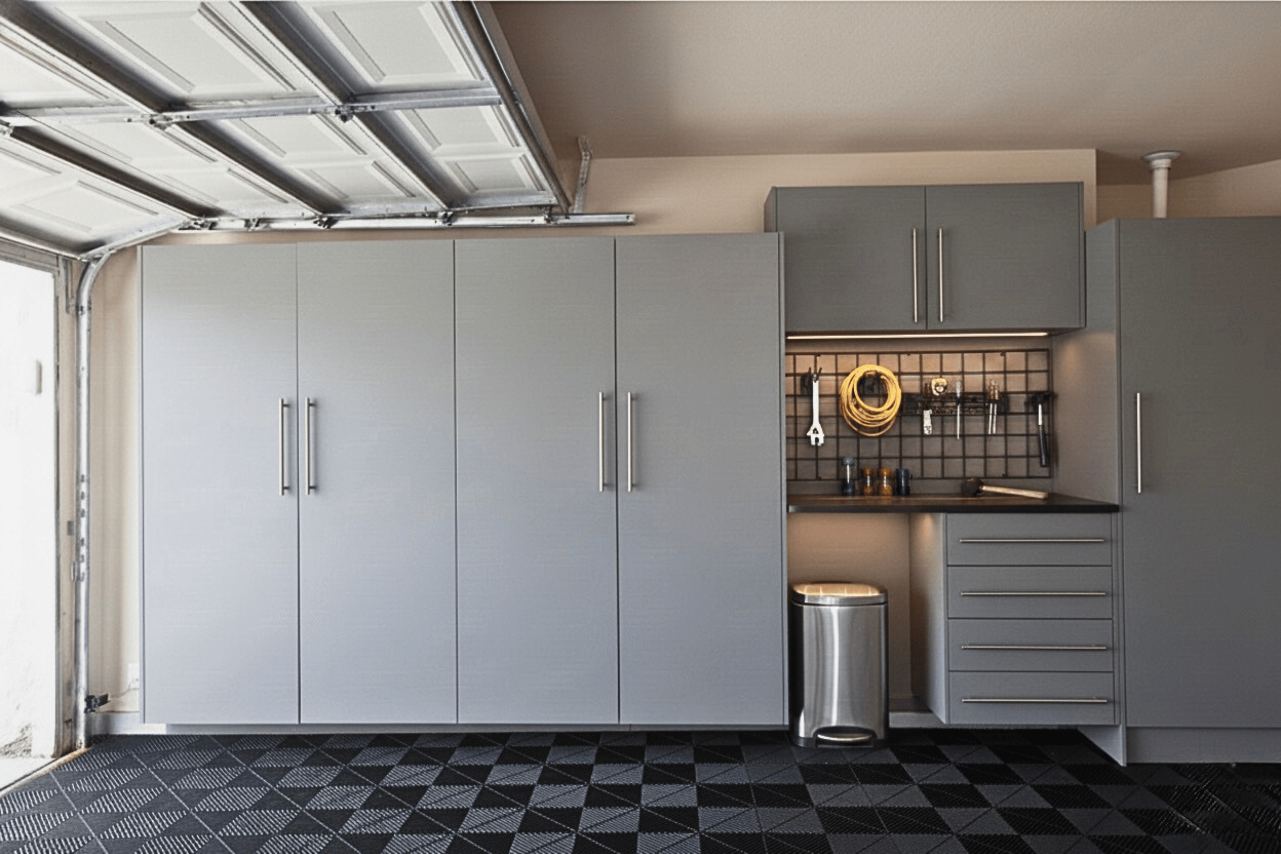 Gray garage cabinets and a work station with hooks, a trash bin, and a patterned black and gray floor.