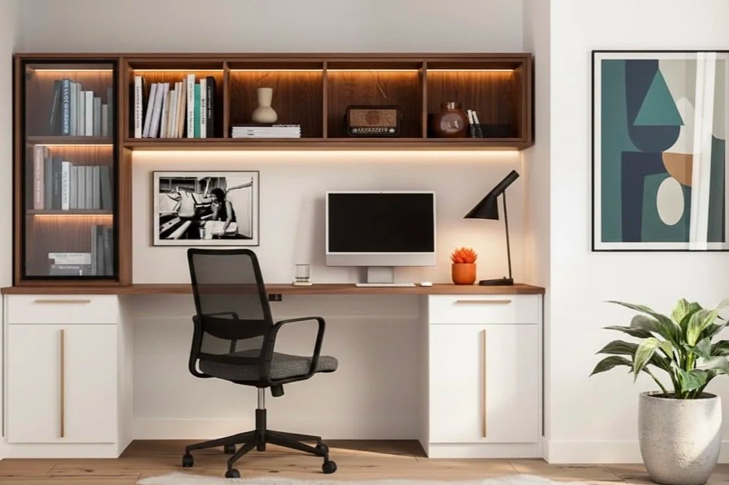 Modern home office with a built-in desk, black ergonomic chair, desktop computer, table lamp, potted plant, decorative framed artwork, and bookshelf with books and decorative items.