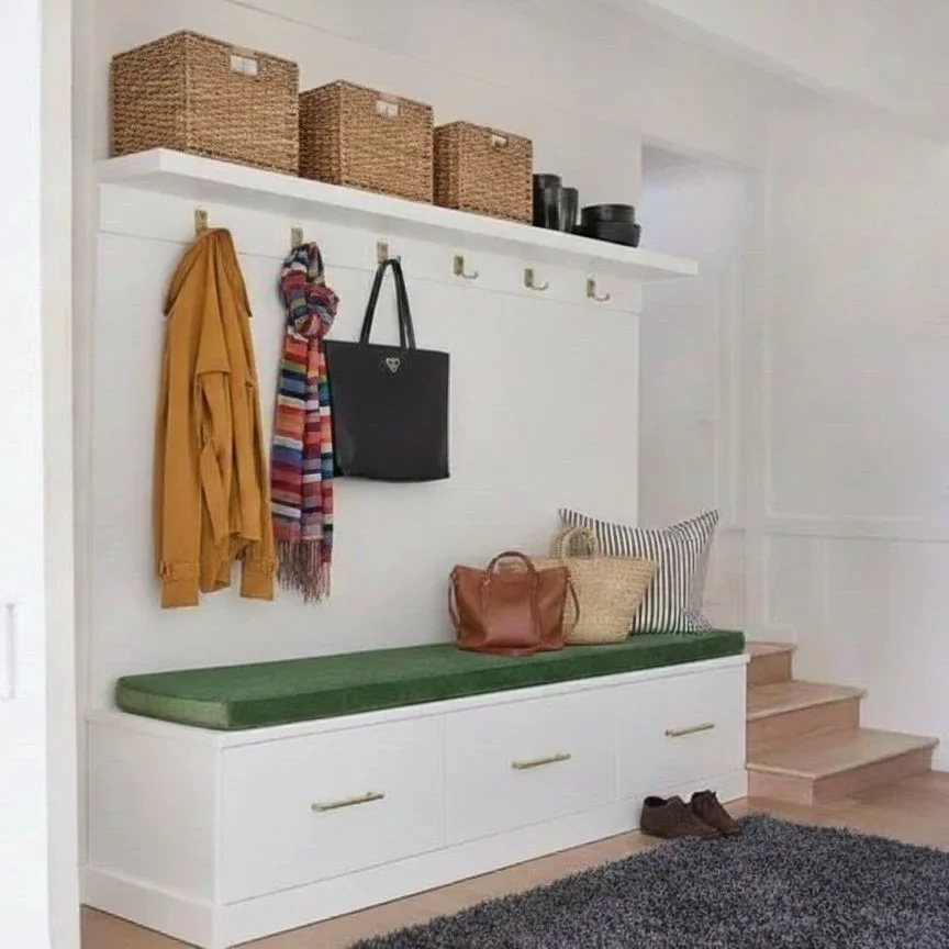 Entryway with a white built-in bench, green cushion, and storage drawers, overhead wooden hooks with items hanging, a striped pillow, a brown purse, a black tote bag, and a woven basket, with a staircase to the right and woven baskets on a shelf abov