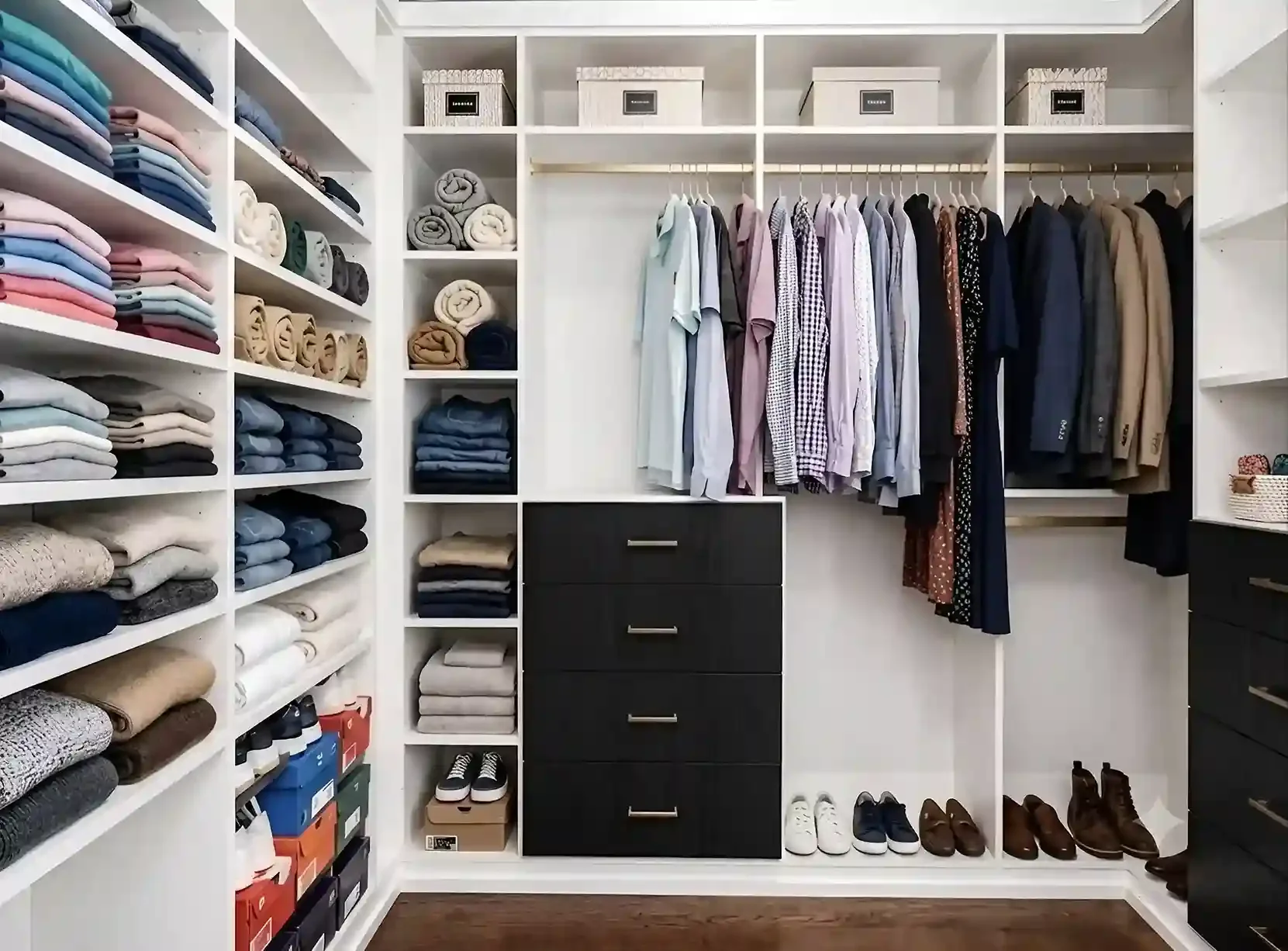 A neatly organized walk-in closet with shelves of folded clothes, boxes, shoes, and hanging garments including shirts, jackets, and suits.