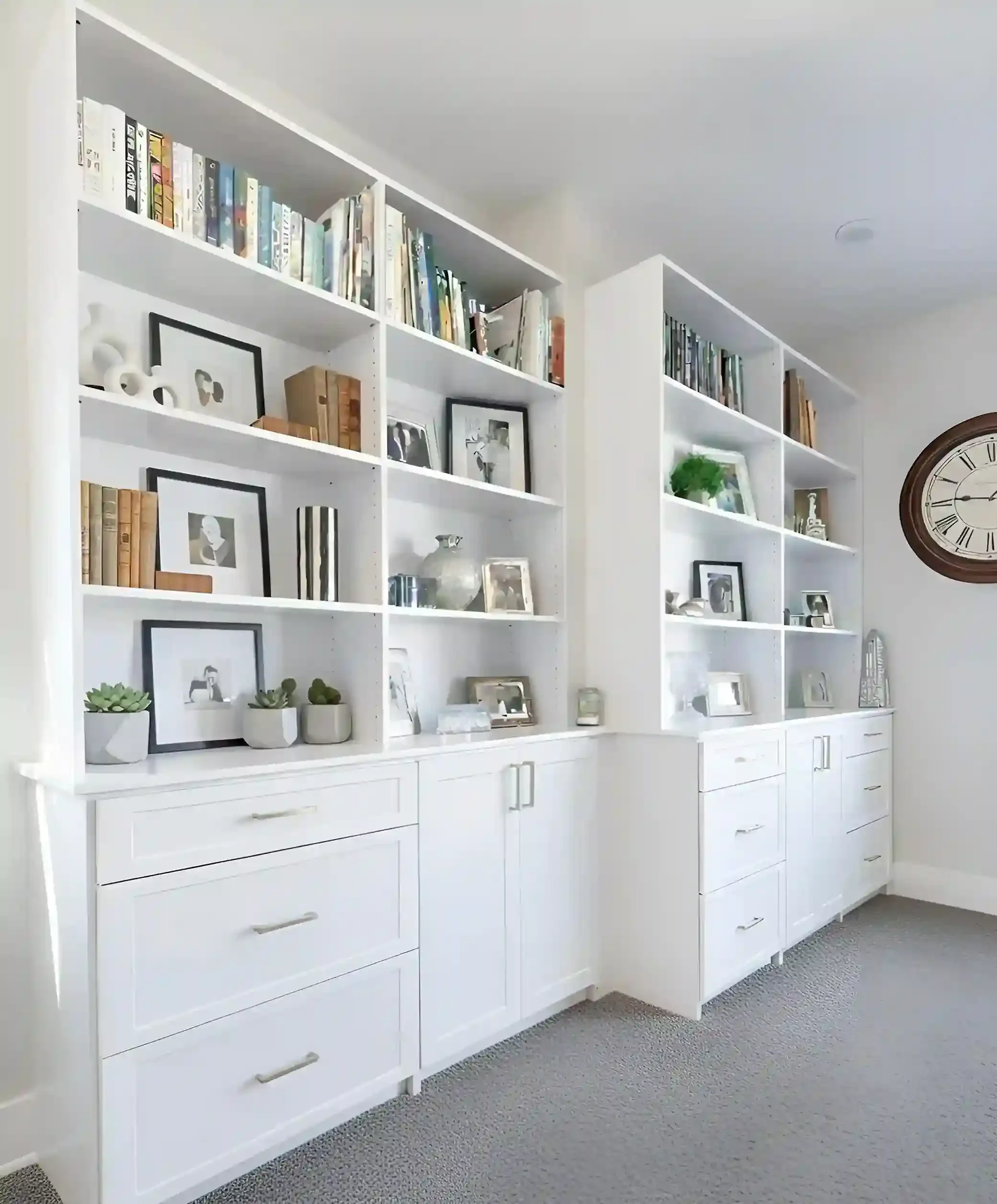 White built-in bookshelves filled with books, framed photos, and decorative items in a living room with beige carpet and a large wall clock.