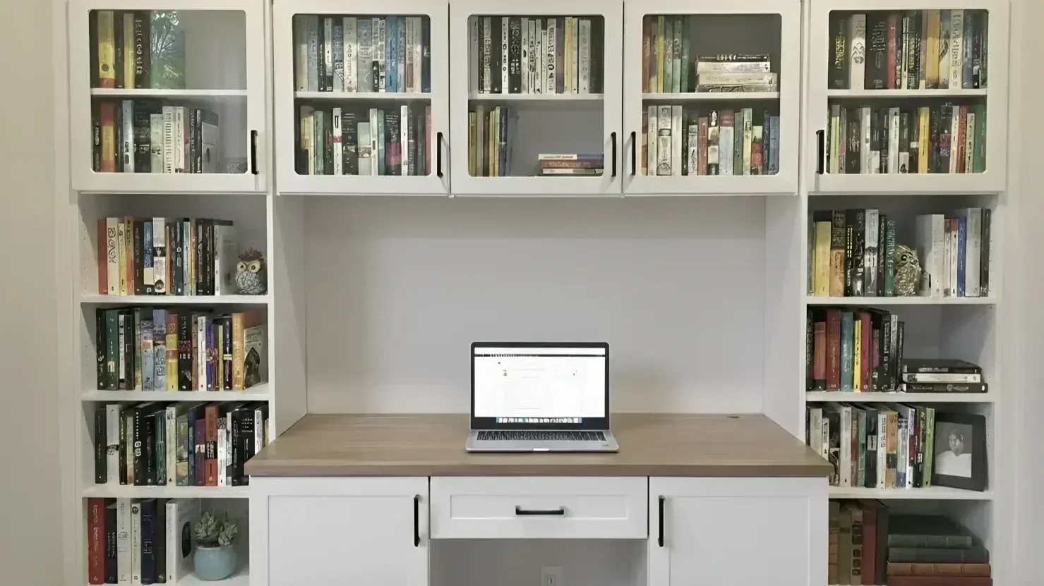 White bookshelf with glass doors filled with books and decorative items, with a wooden desk in front holding a laptop.