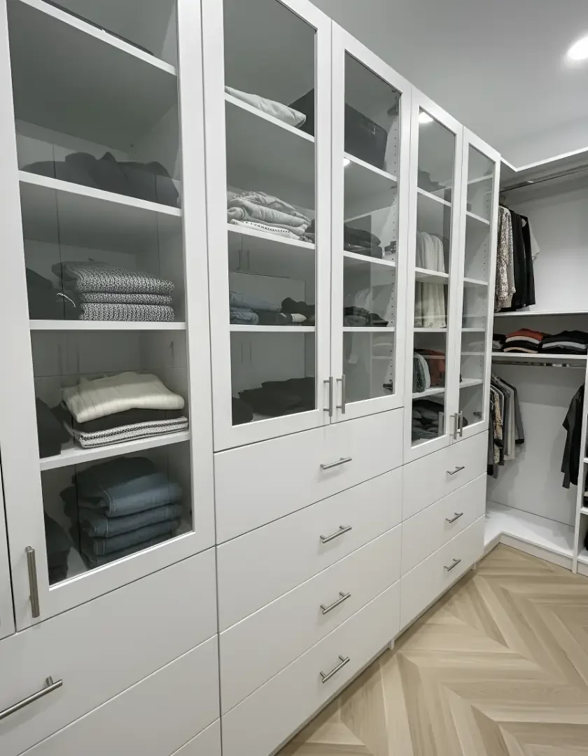 Closet with white drawers and glass-front cabinets filled with folded clothes, with more clothing hanging in the background.