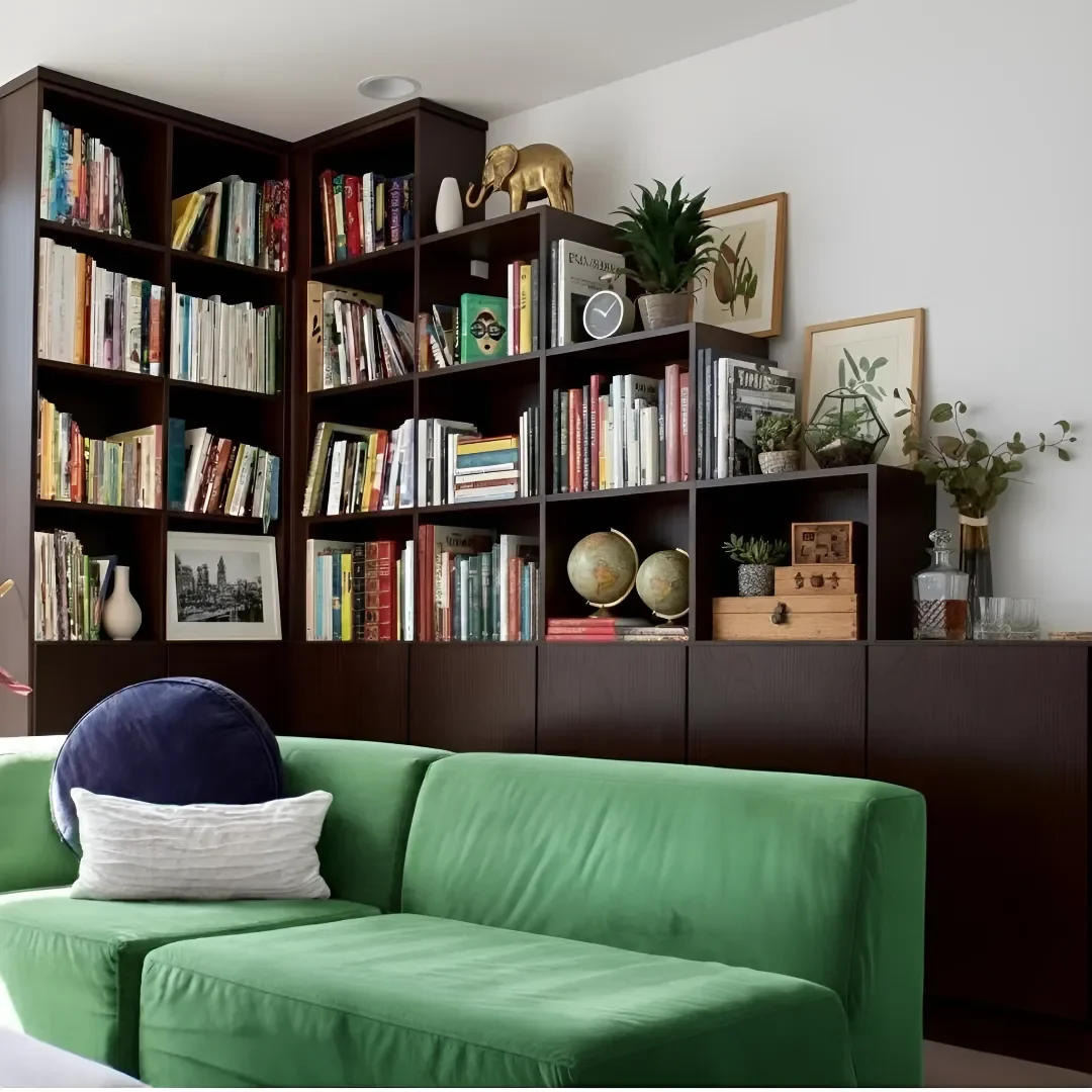 A green sofa with a white pillow and a dark blue headrest in front of a dark wooden bookshelf filled with books, decorative items, plants, and framed art.