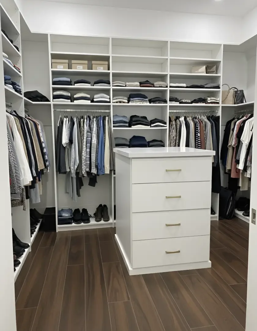 A walk-in closet with white shelves filled with folded clothes, hanging shirts, and shoes on the floor. A white chest with four drawers is in the center.