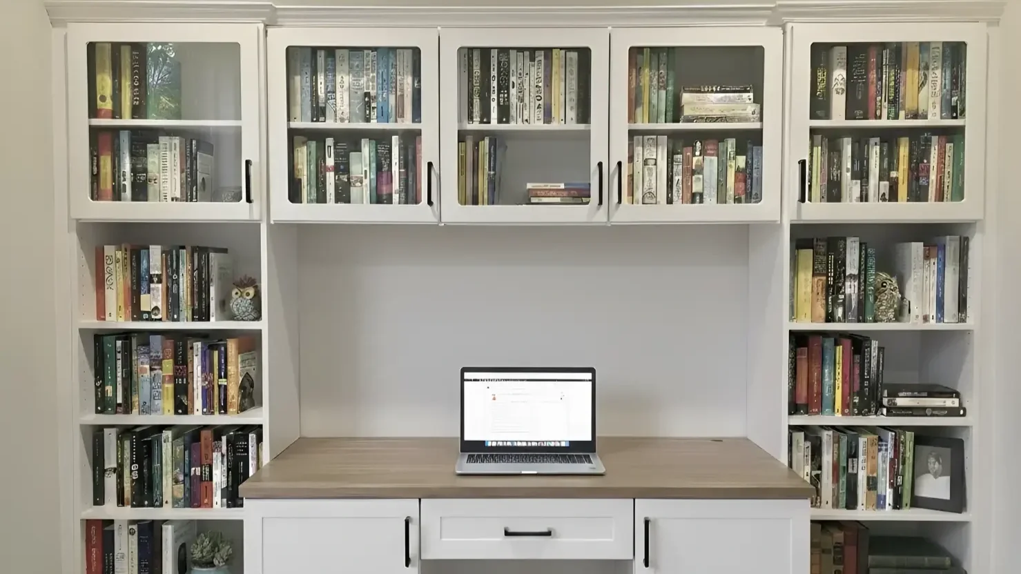 White bookshelf with glass cabinets on top filled with books, a wooden desk surface with a laptop, and shelves with books and decorative items on either side.