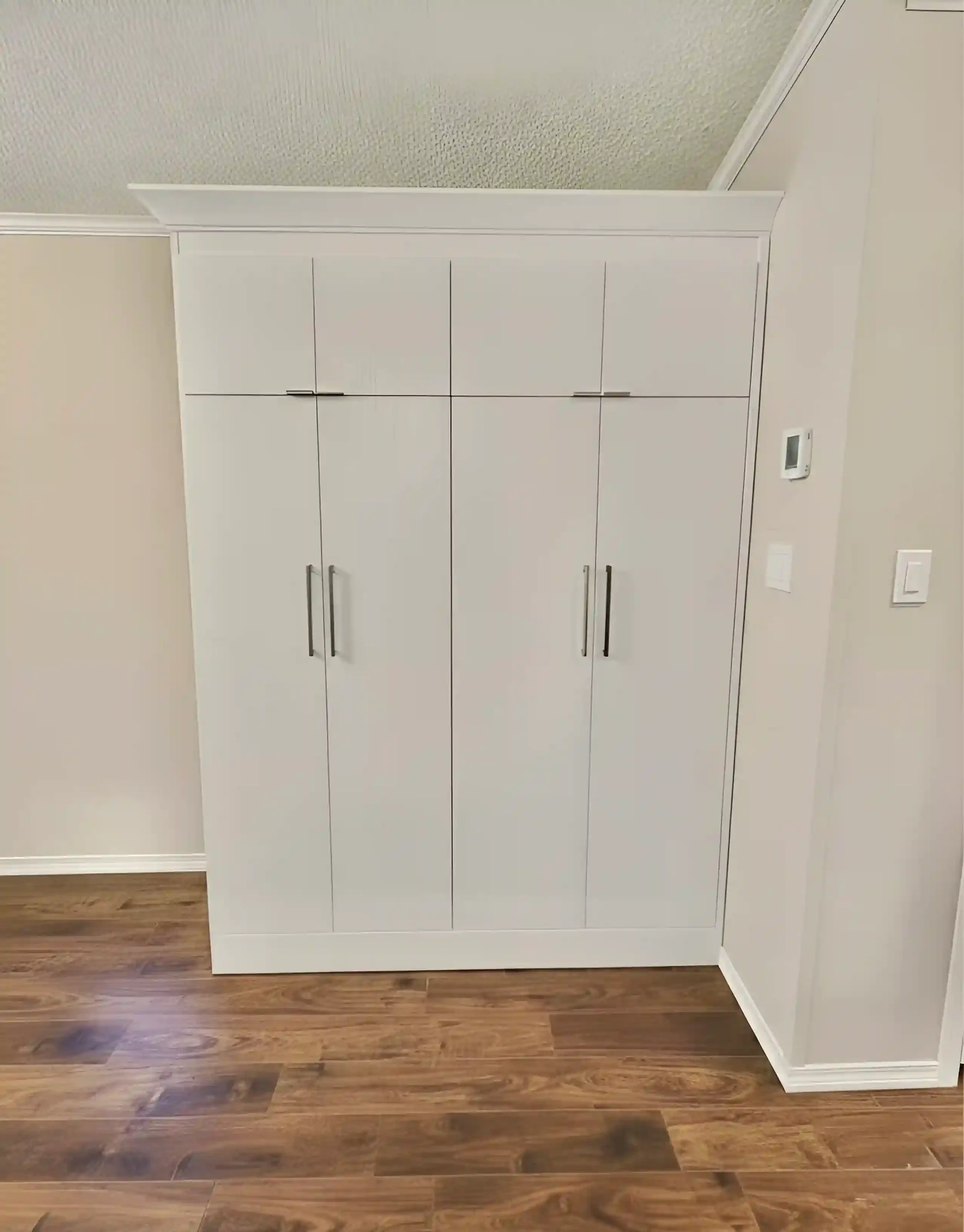 White built-in closet with multiple doors and silver handles in a room with wood flooring and beige walls.