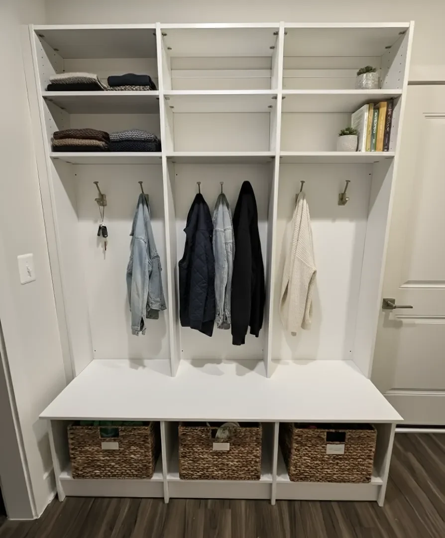 A white entryway bench with storage baskets underneath and hooks on the wall for hanging coats, scarves, or keys. Some folded clothes and decorative items are on the shelves above.