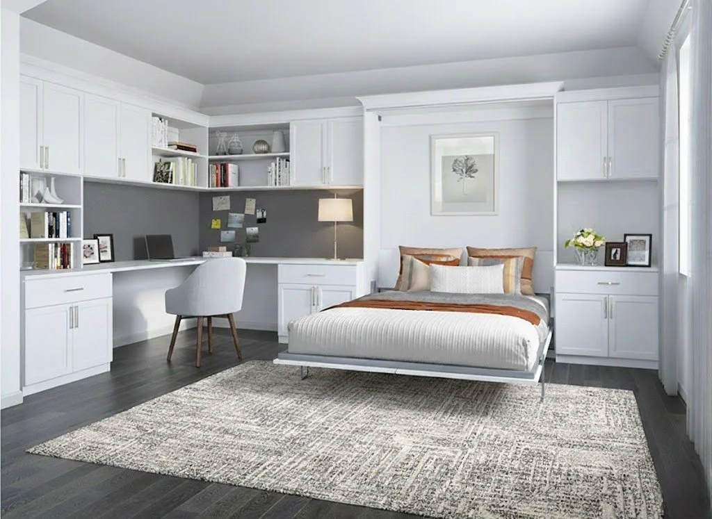 A modern bedroom with a white bed, built-in white cabinets and shelves, a desk with a chair, a table lamp, framed artwork, a flower arrangement, and a beige rug on dark wood floors.