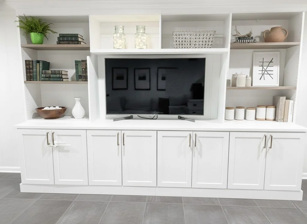 White entertainment center with shelves, a flat-screen TV, decorative books, and various decorative items including jars, framed art, and plants.