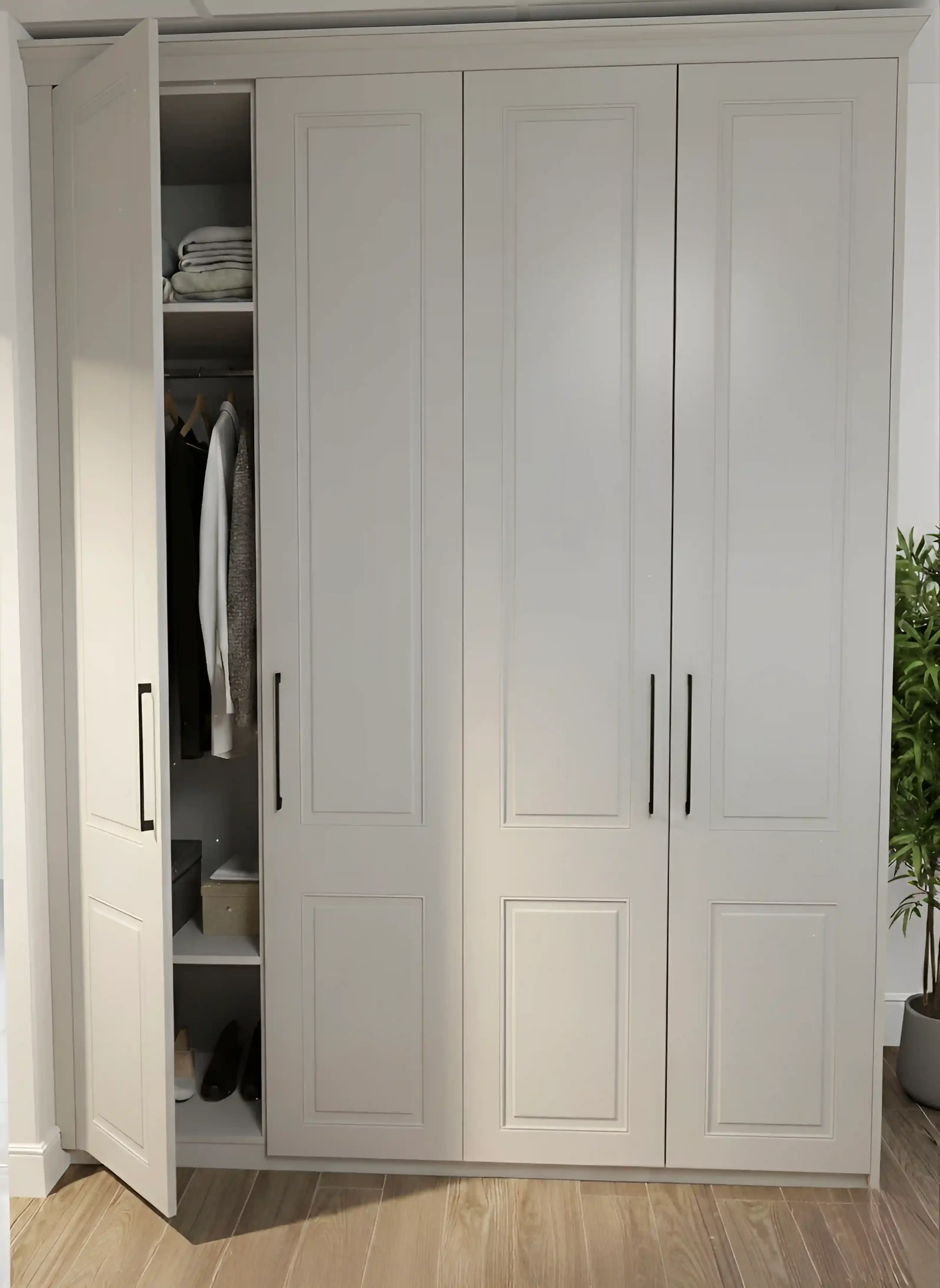 A white wardrobe with one door open, revealing shelves and hanging clothes inside, next to a potted plant and wooden flooring.