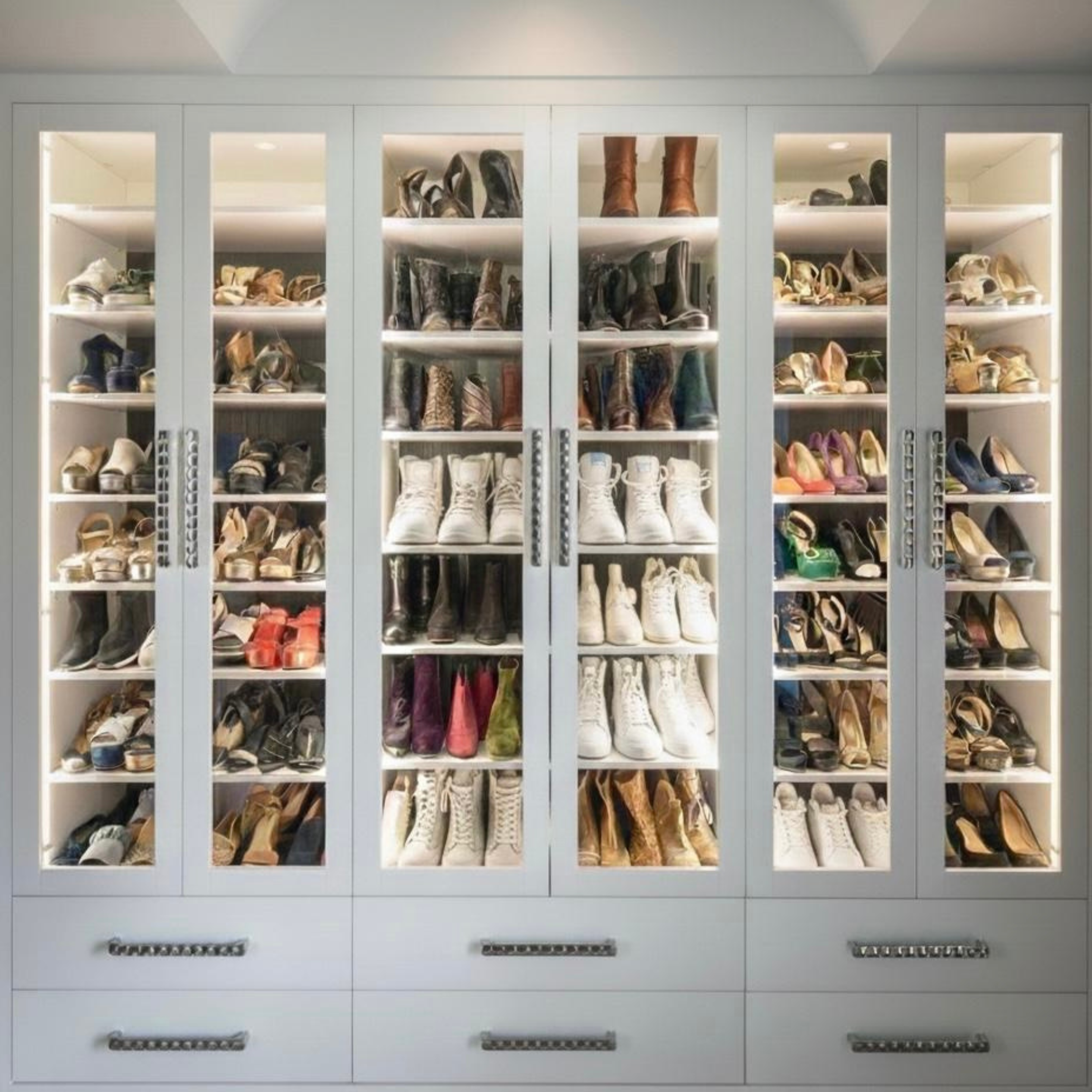 A white shoe cabinet with glass doors filled with neatly organized shoes and boots on shelves, with drawers at the bottom.