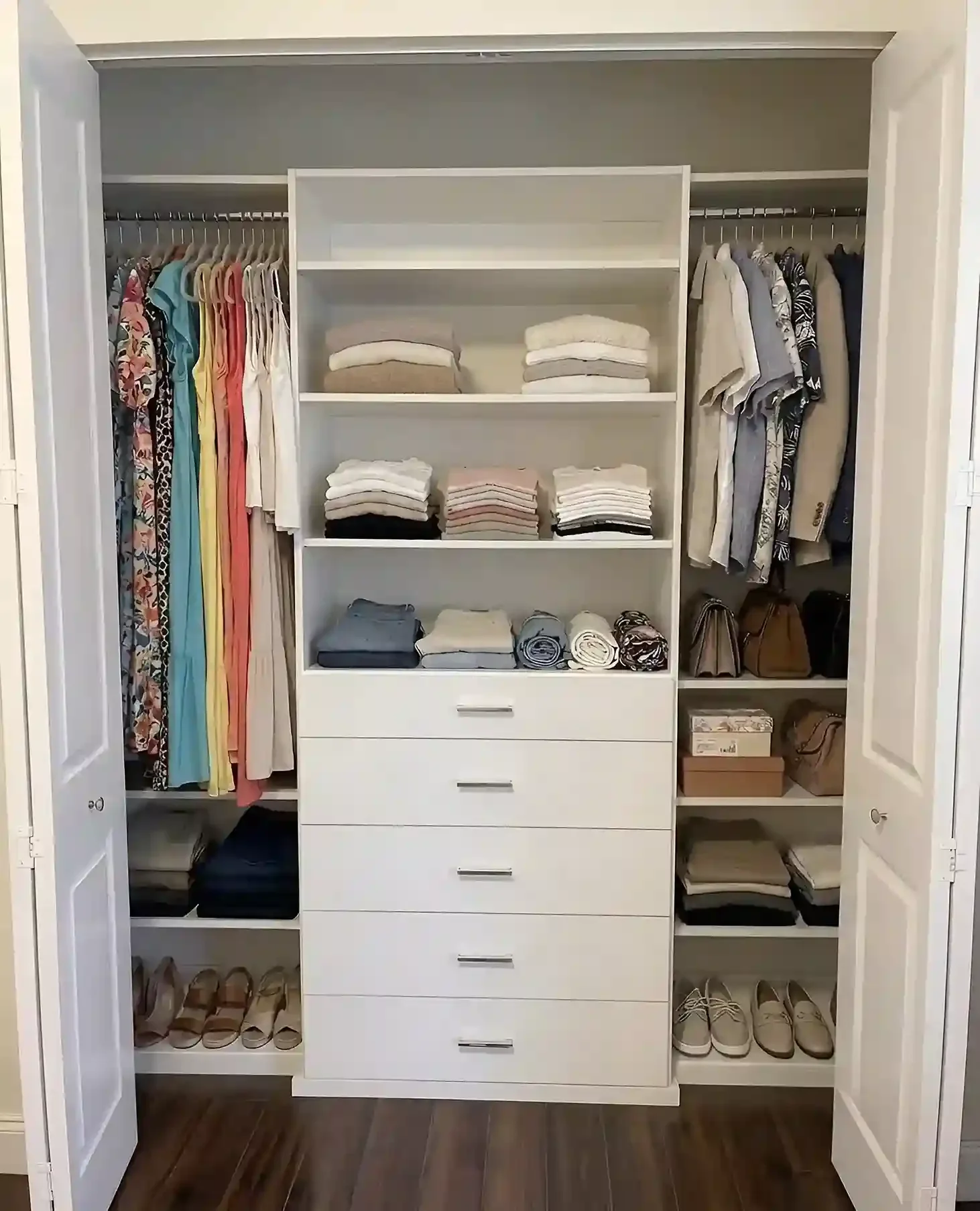 An organized closet with open white doors. Inside, there is a mixture of tank tops, shirts, folded towels, and shoes on shelves, along with handbags and boxes.