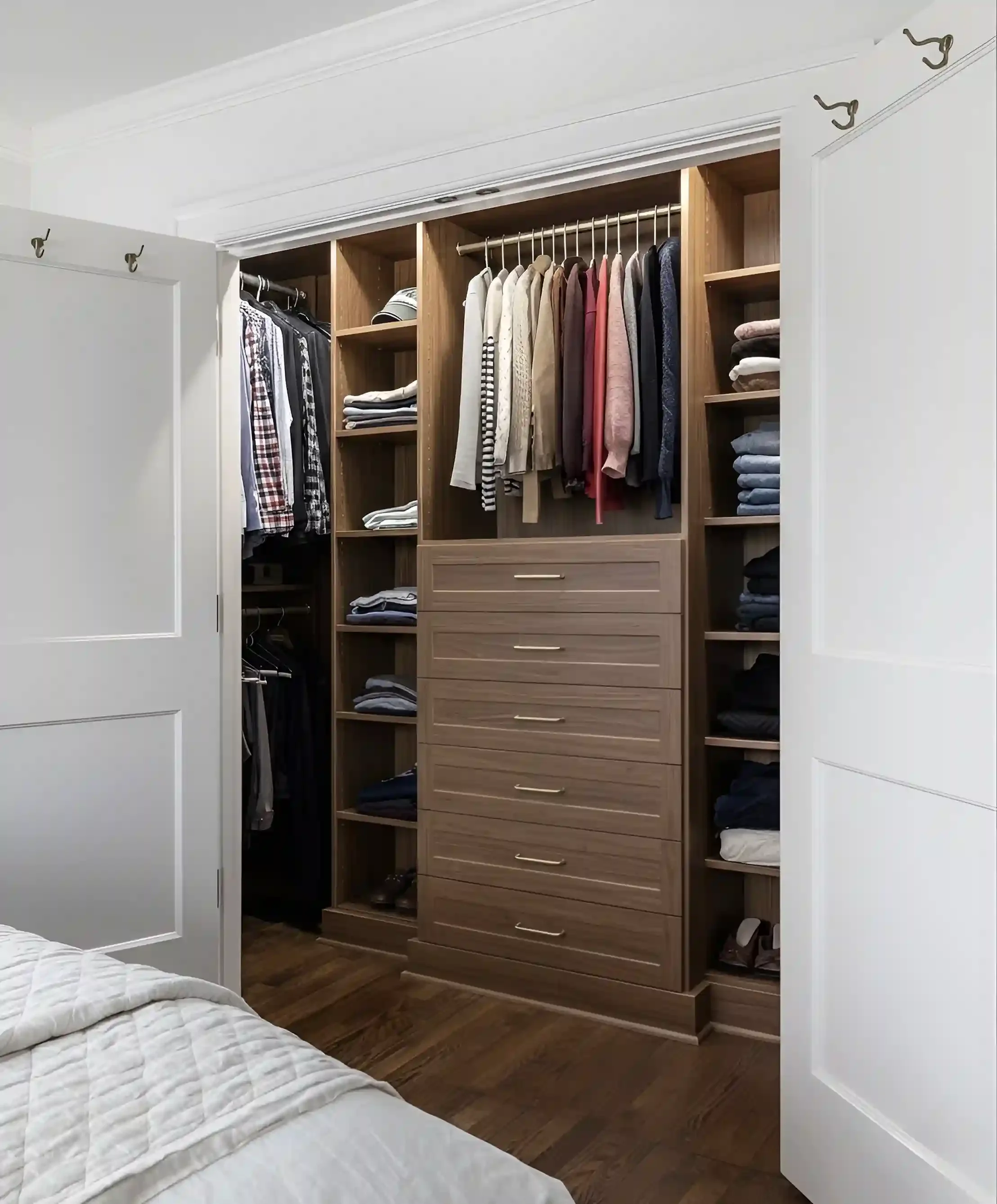 Open closet with dresser and shelves filled with folded clothes, hanging shirts, and pants, in a room with hardwood floors and white walls.