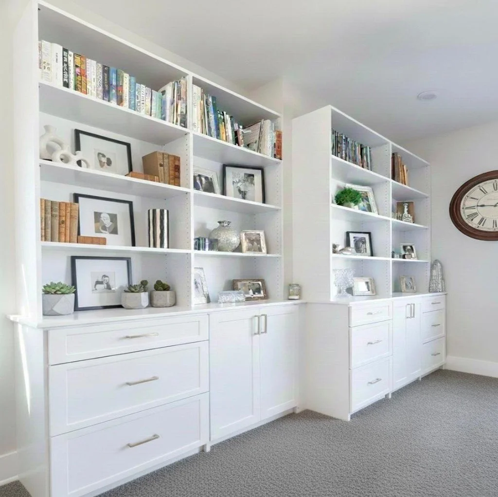 White built-in bookshelves filled with books, framed photos, decorative objects, and small potted plants in a living room.