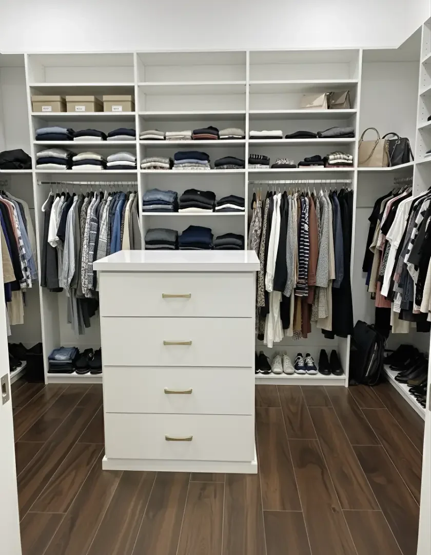 A walk-in closet with white shelves filled with folded clothes, hanging shirts, pants, and accessories, and a white chest of drawers in the center on wood flooring.
